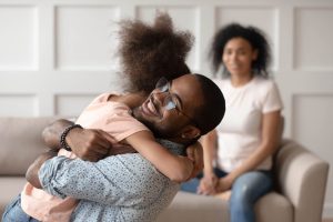 תמונת נושא של המאמר על משמורת משותפת מתחת לגיל 6 המציגה אב מחבק בחום את בתו הצעירה על ספה בסלון מואר, שניהם מחייכים. האב מרכיב משקפיים עגולים ומשופעים. האם יושבת מטושטשת ברקע.