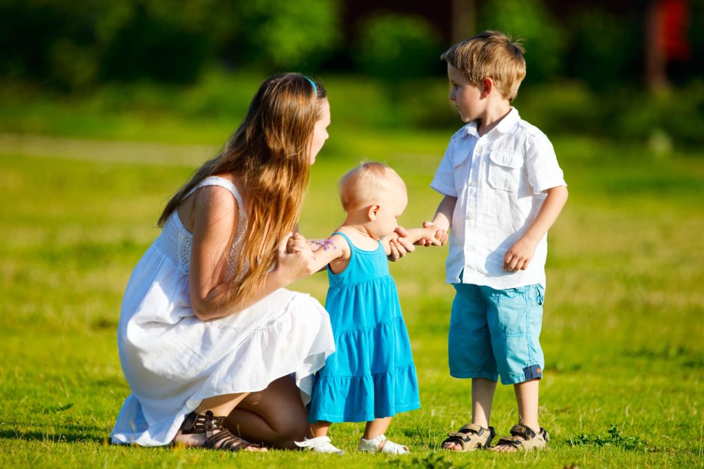 תמונת נושא של המאמר על הסכם הורות משותפת המצגיה אישה בלונדינית על דשא ירוק לובשת שמלה לבנה עם שני ילדים הילדים לבושים בכחול ולבן התינוקת אוחזת בידה של האישה ובן שתי עומד בצד.