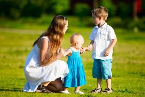 תמונת נושא של המאמר על הסכם הורות משותפת המצגיה אישה בלונדינית על דשא ירוק לובשת שמלה לבנה עם שני ילדים הילדים לבושים בכחול ולבן התינוקת אוחזת בידה של האישה ובן שתי עומד בצד.